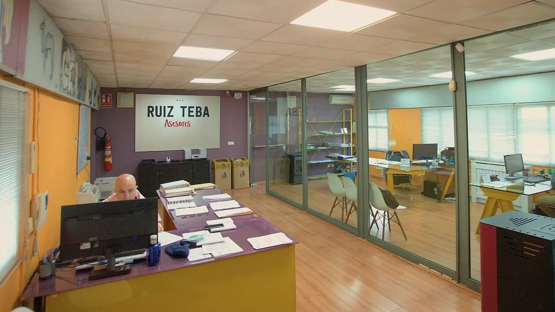 Interior del despacho de Ruiz Teba Asesores en Torrejón de Ardoz: el rótulo de la marca en la pared, un puesto de trabajo con expedientes y los despachos acristalados al fondo